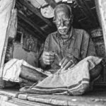 grayscale photo of man sewing textile while sitting on floor